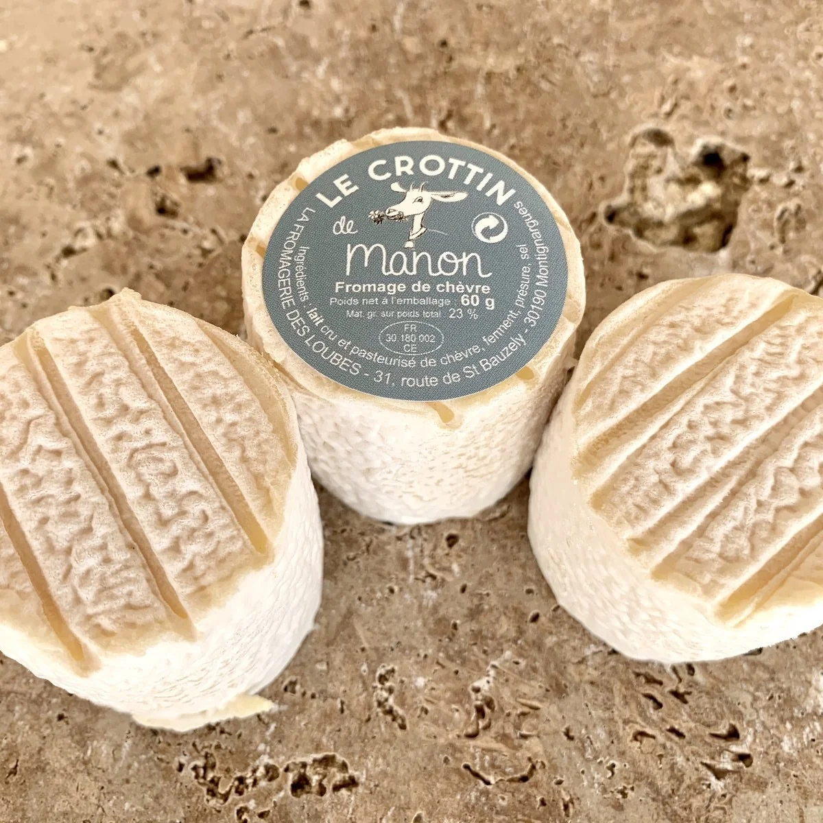 Trois Crottins de Manon sur une pierre ; celui du milieu porte l'étiquette « Le Crottin de Manon » de la Fromagerie des Loubes